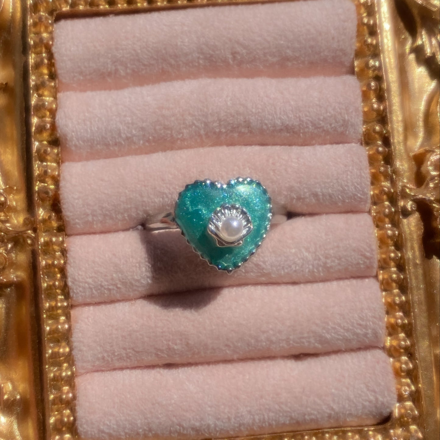 A heart-shaped turquoise ring with a central white pearl surrounded by a halo of small clear stones, set on a silver band. The ring is displayed on a light pink velvet ring roll inside an ornate gold frame.