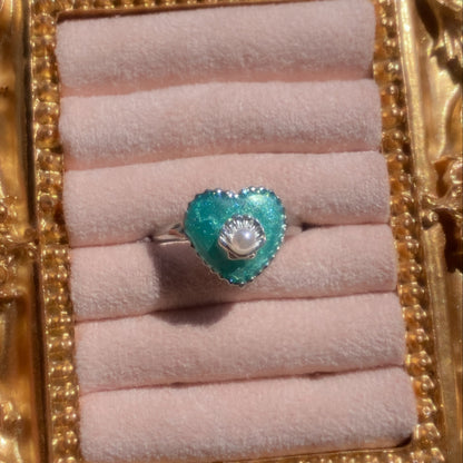A heart-shaped turquoise ring with a central white pearl surrounded by a halo of small clear stones, set on a silver band. The ring is displayed on a light pink velvet ring roll inside an ornate gold frame.