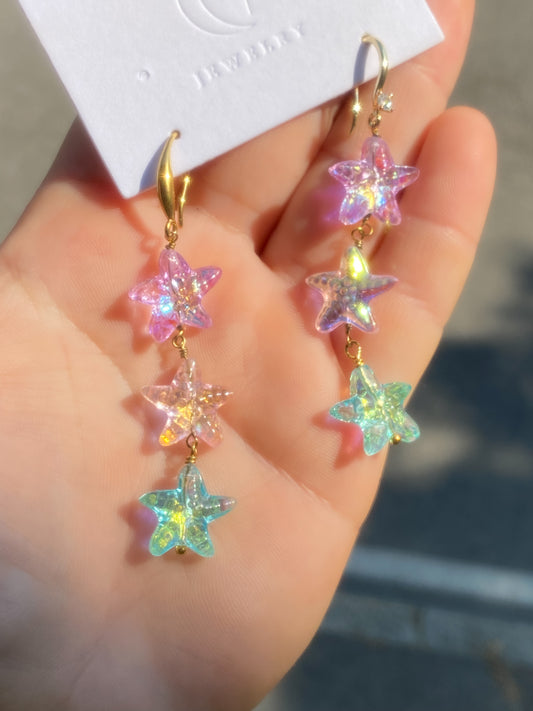 A pair of dangling star-shaped earrings held in a hand. Each earring has gold-tone hooks and three iridescent pastel stars in pink, yellow, and mint green, attached to a white jewelry card.ish