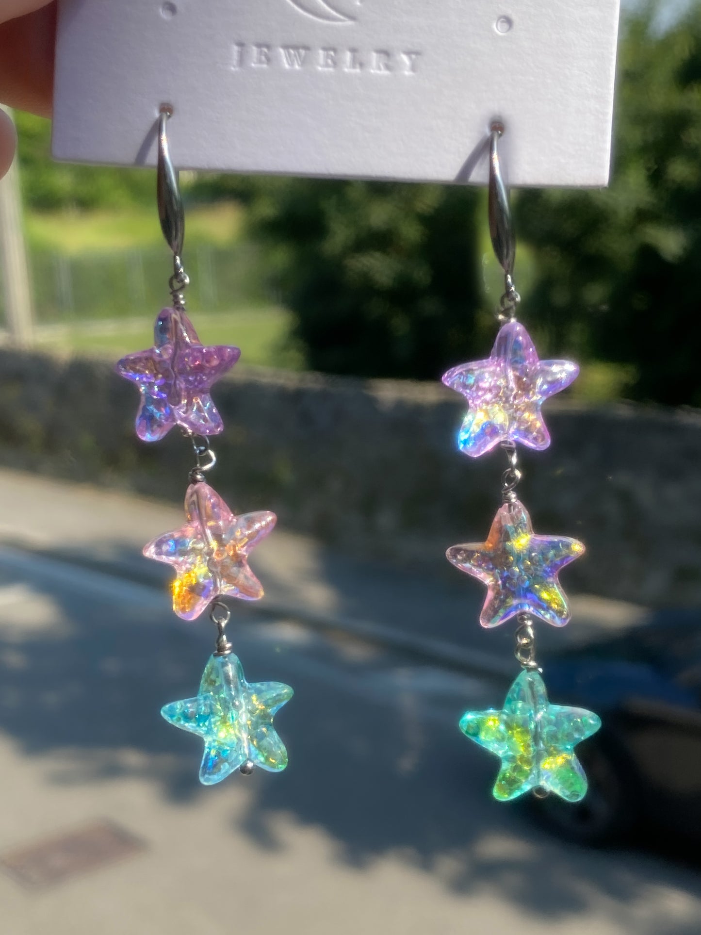 A pair of dangling starfish-shaped earrings with iridescent, holographic colors, hanging from a white jewelry display card held by a hand. The background is a blurred outdoor scene with green foliage and a stone wall.